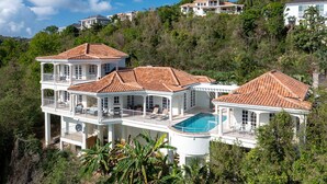 A large, multi-story house with a swimming pool, surrounded by lush greenery and other houses on a hillside. - Bananaquit Villa: Amazing Views! Large Pool! Full AC! (St John)