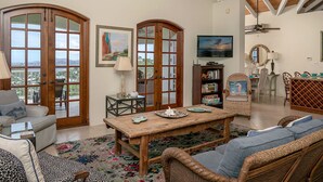 A spacious living room with a wooden table, wicker furniture, and large windows with wooden frames. - Bananaquit Villa: Amazing Views! Large Pool! Full AC! (St John)