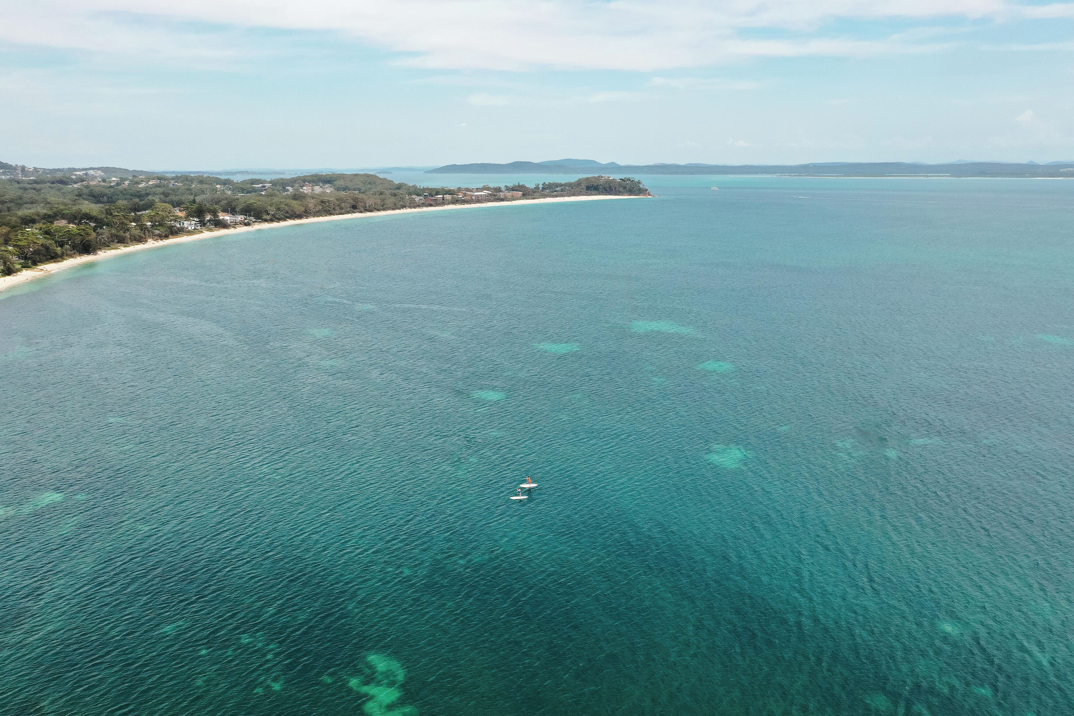 An aerial view of a coastal area with clear turquoise waters, a sandy beach, and a small boat in the distance.