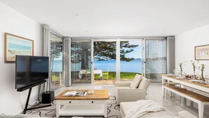 A modern living room with a large TV, a wooden coffee table, a sofa, and a dining area with a view of the sea. - Tranquil Waters - Luxury Waterfront Home  with Stunning Views (Salamander Bay)