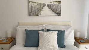 A neatly made bed with white and blue pillows, a framed beach painting above it, and a wooden nightstand with a lamp and a book. - Three Views Lodge (Torpoint)