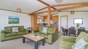 A spacious living area with green sofas, a wooden coffee table, and a dining table with chairs. A kitchen is visible in the background. - Three Views Lodge (Torpoint)