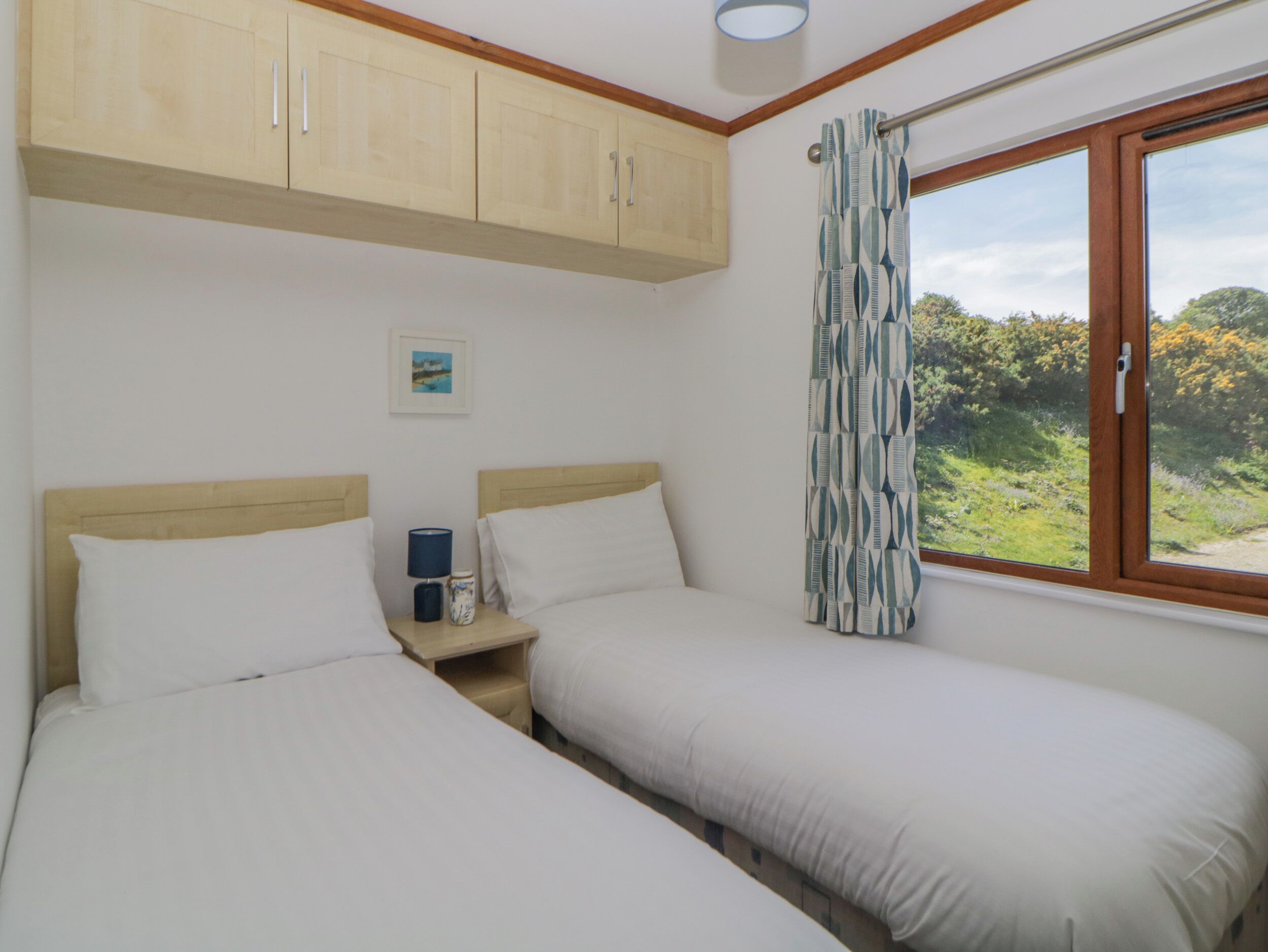 A bedroom with two single beds, wooden cabinets above, a small bedside table with a lamp, and a window with a curtain.