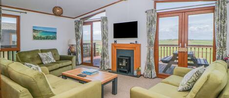 A living room with green sofas, a fireplace, a flat-screen TV, and large windows with curtains.