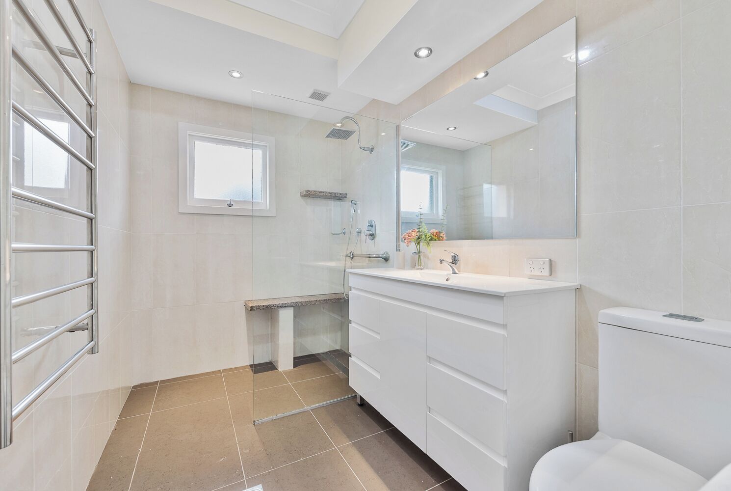 A modern bathroom with a glass shower enclosure, a large mirror, and a white sink with a vase of flowers.