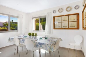 A dining area with a table set for four, white chairs, a window with curtains, and a wall with framed nautical artwork. - Villa Estanyol (Campos)