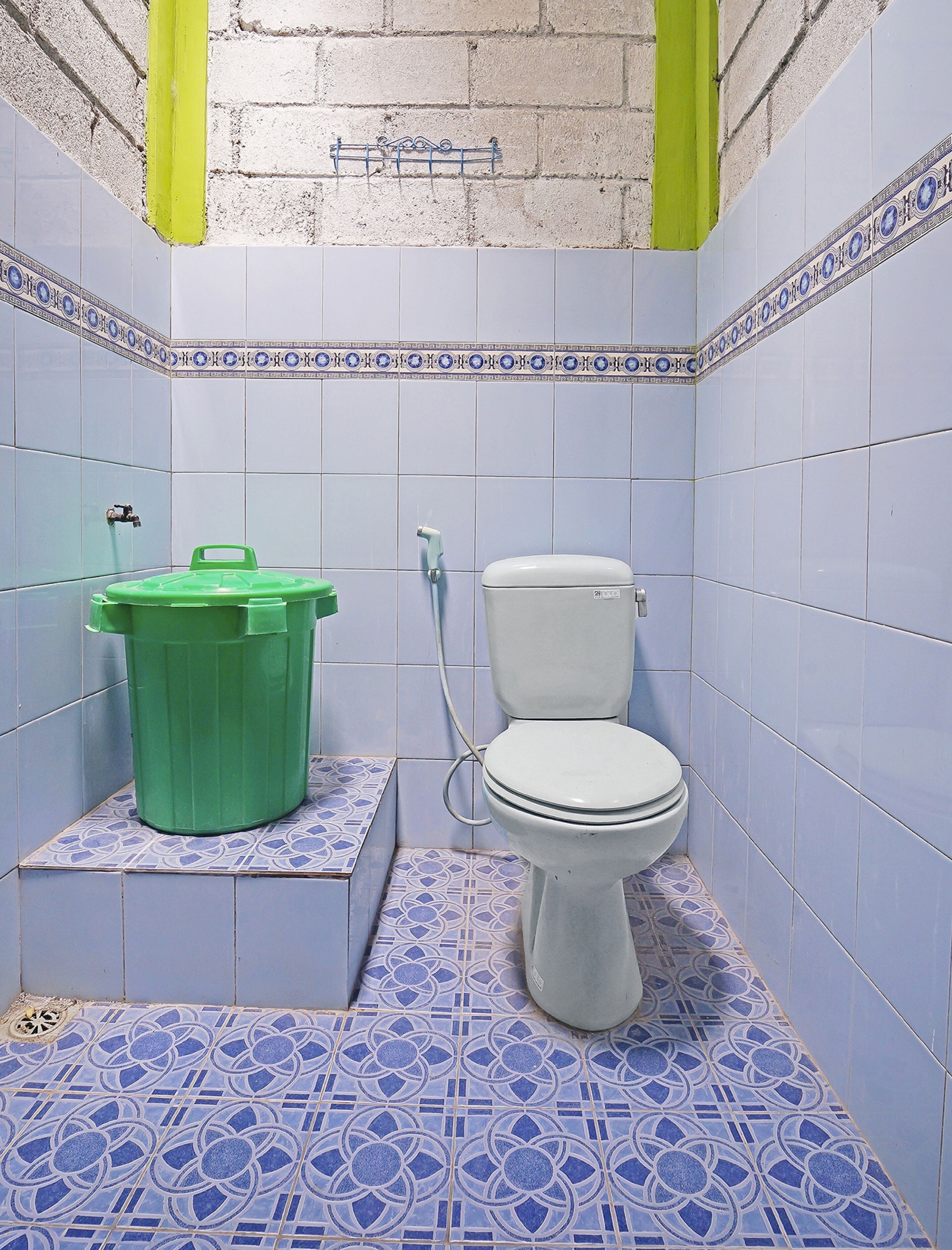 A small bathroom with a toilet, a green trash bin, and a shower hose.