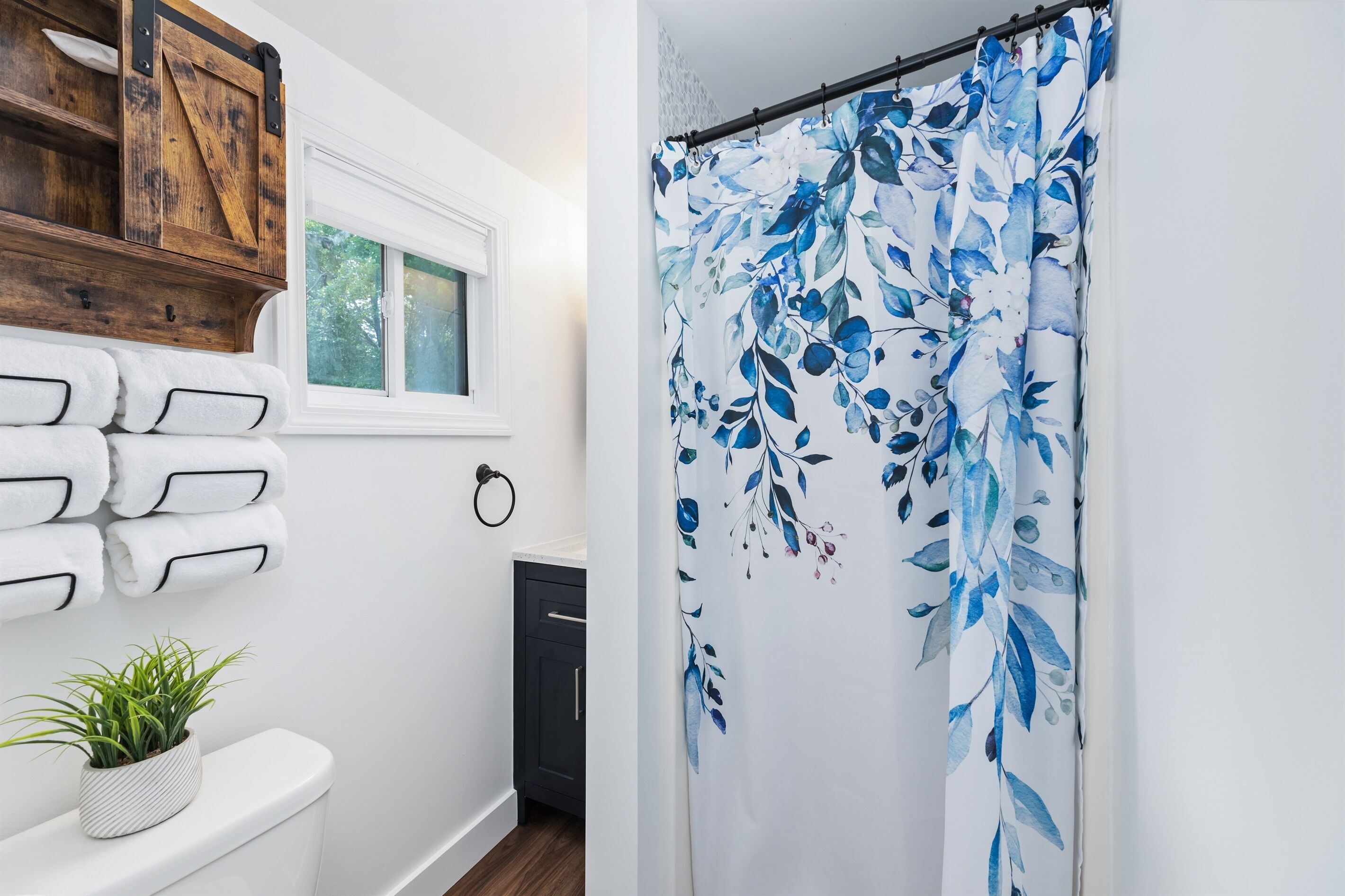 Een badkamer met witte muren, een houten plank met handdoeken, een plant in pot en een douchegordijn met een blauw bloemenmotief.