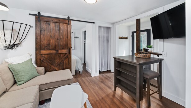 A modern living room with a wooden door, a sofa, a dining table with chairs, and a mounted TV.