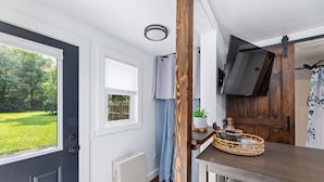 A room with a dark door, a window with a view of greenery, a ceiling light, a wooden wall, a TV mounted on the wall, a small table with a basket, and a radiator.