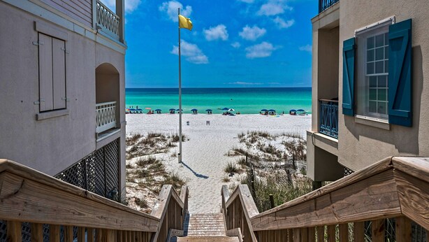 Vista de una playa desde un edificio con asta de bandera, escaleras de madera y una playa arenosa con sombrillas.