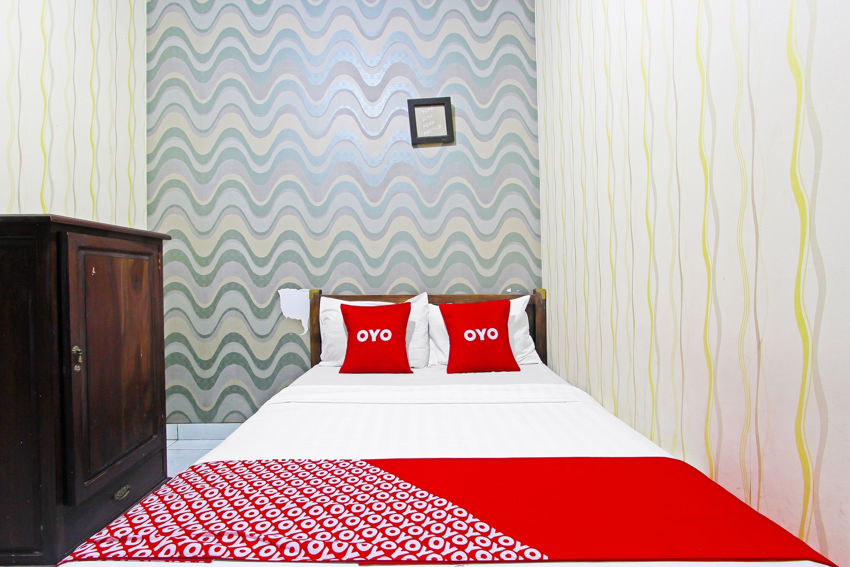 Une chambre avec un lit, deux oreillers rouges ornés du texte « OYO », une armoire en bois et un papier peint à motifs.