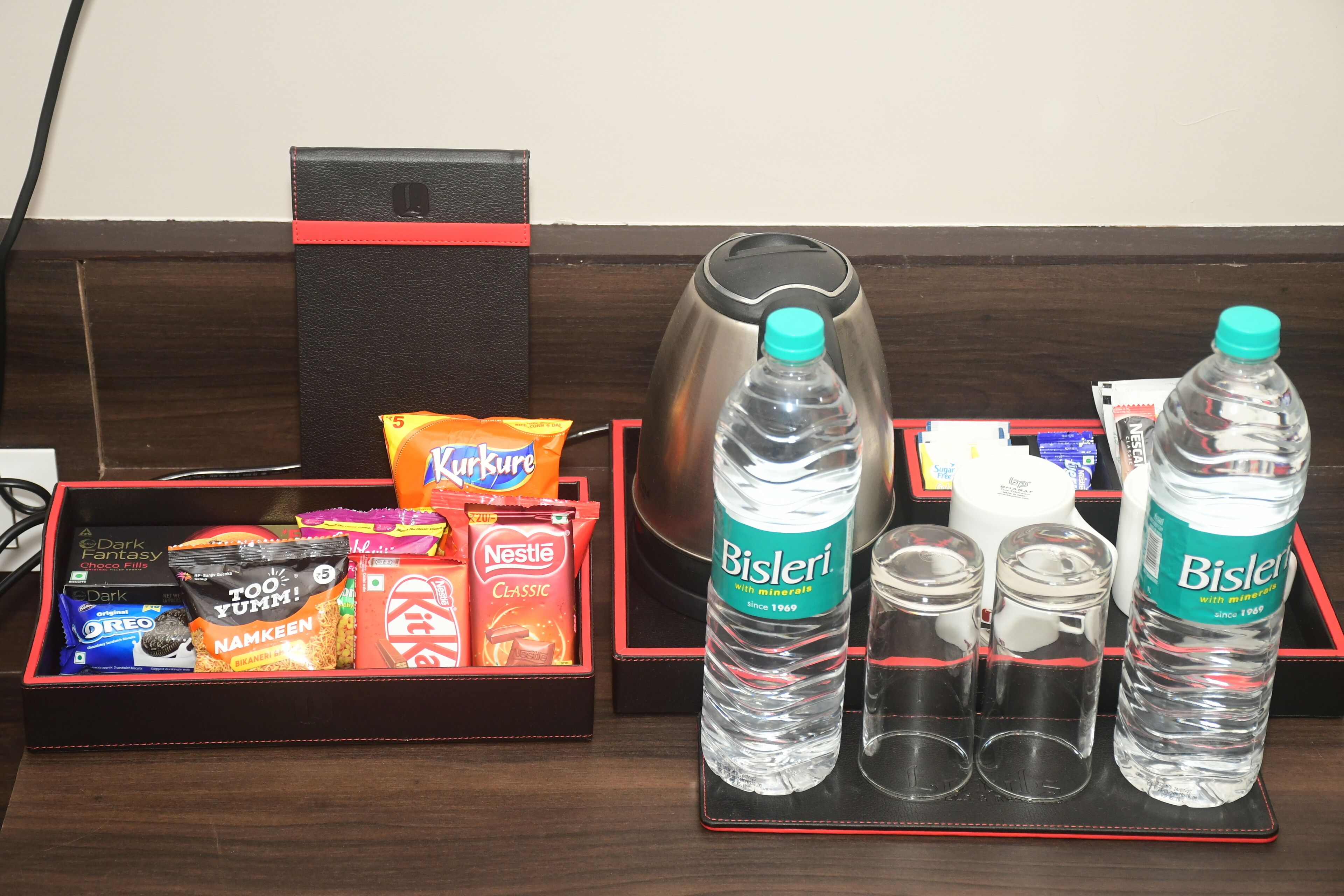 A hotel reception area with a black leather desk, a red and black tray with snacks, Bisleri water bottles, and a coffee maker.