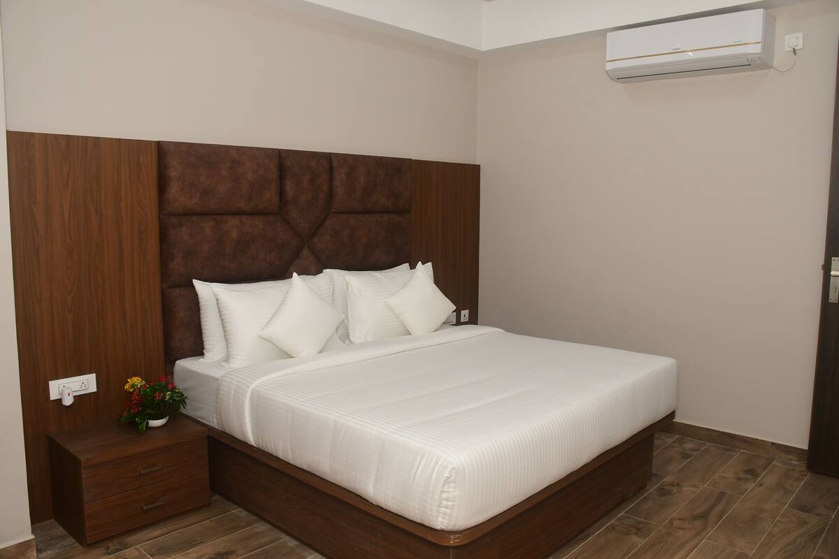 A bedroom with a wooden headboard, a white bedspread, a nightstand with a plant, and an air conditioning unit on the wall.