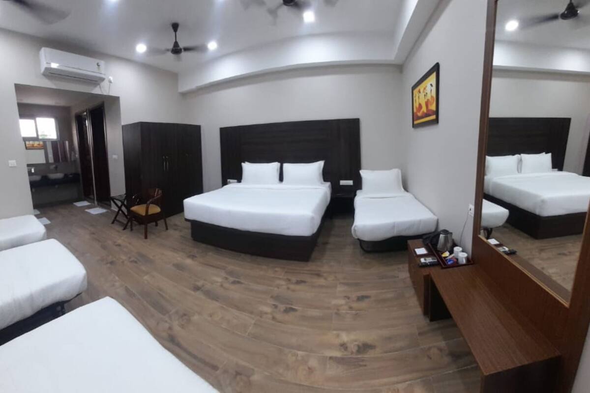 A hotel room with two single beds, a double bed, a wooden floor, a ceiling fan, an air conditioning unit, a small table with a chair, and a mirror.
