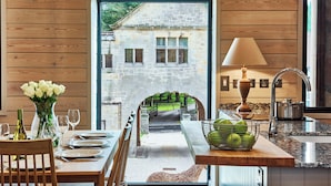 Un coin repas offrant une vue sur un bâtiment en pierre, visible à travers une arche. On y aperçoit une table dressée avec des assiettes et des verres, un vase rempli de fleurs et un panier de pommes vertes.