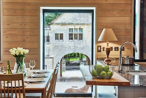 A dining area with a view of a stone building through an archway, a table set with plates and glasses, a vase of flowers, and a basket of green apples.