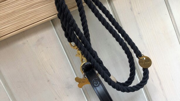 A black braided necklace with a metal pendant and a wooden hook on a white wooden surface. - The Carriage House (Hexham)