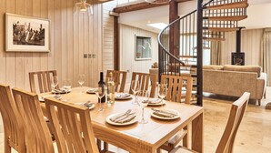 A dining area with a wooden table set for six, surrounded by wooden chairs. A spiral staircase is visible in the background, leading to an upper level. - The Carriage House (Hexham)
