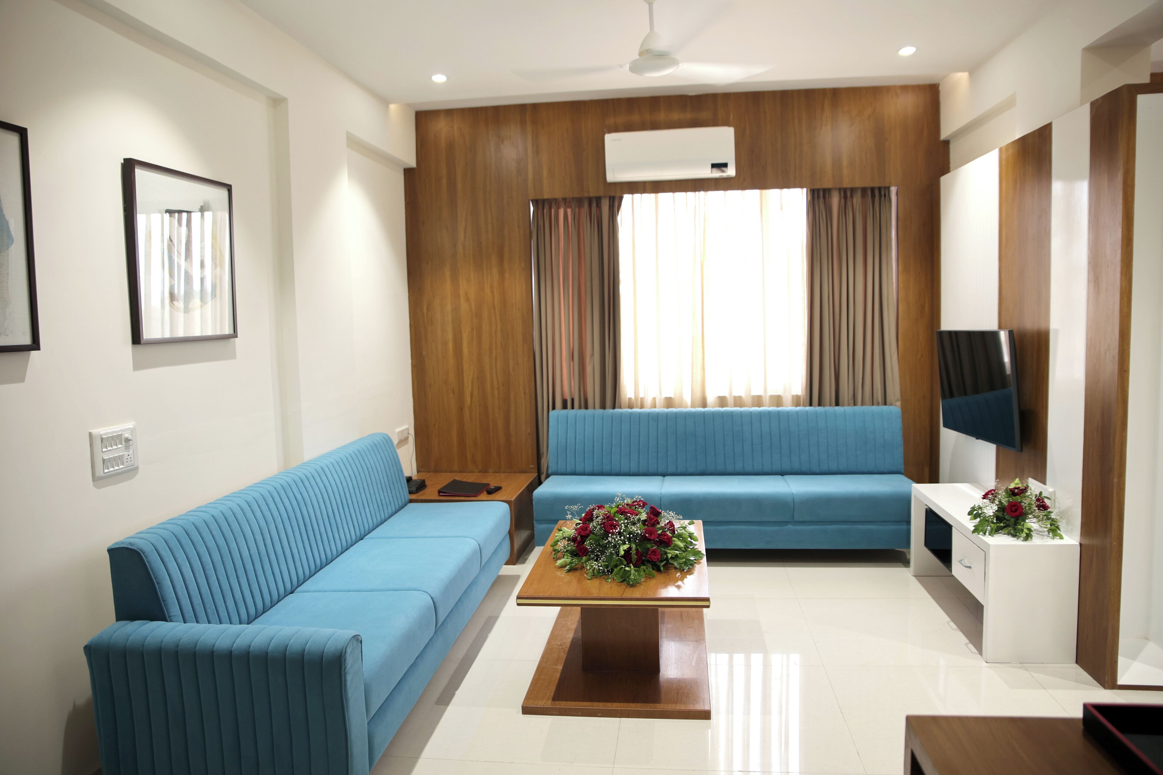 Una sala moderna con dos sofás azules, una mesa de centro de madera con un centro de mesa floral, y un televisor de pantalla plana sobre un soporte blanco.