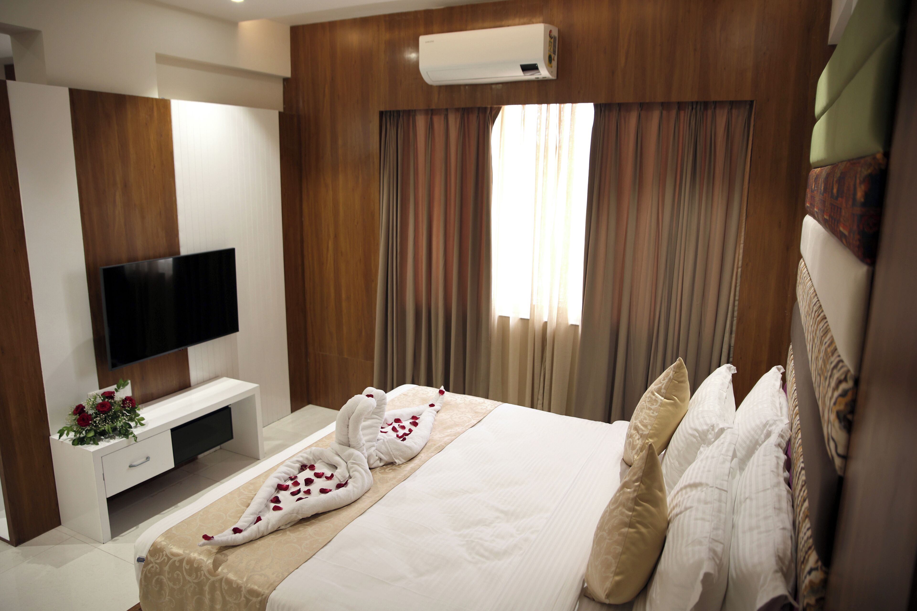Habitación de hotel con televisor de pantalla plana, cama con sábanas blancas, toalla decorativa y ventana con cortinas.