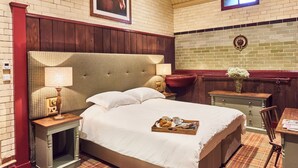 A hotel room with a large bed, a wooden nightstand with a lamp, a small table with a tray of breakfast items, and a brick wall with a framed picture.