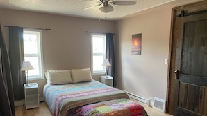 Une chambre à coucher comprenant un lit, deux tables de chevet, un ventilateur de plafond, une fenêtre avec des rideaux, une porte et un radiateur.