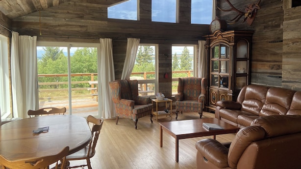 L’intérieur d’un chalet rustique, avec des murs en bois, une table à manger, un canapé en cuir et une grande fenêtre donnant sur une terrasse.