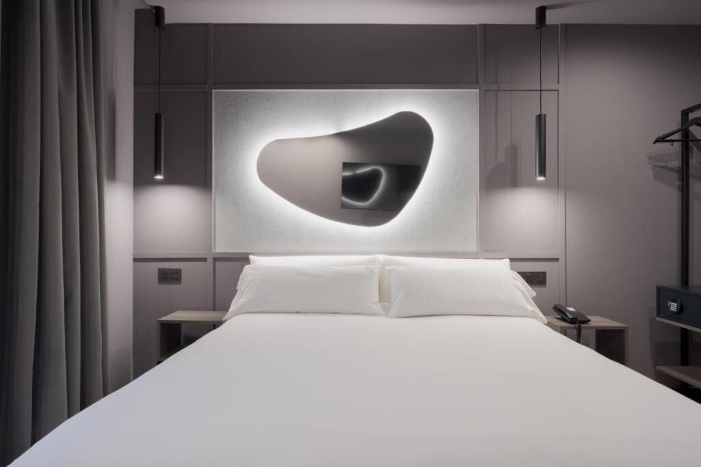 Een moderne hotelkamer met een groot bed, een lamp in de vorm van een hart erboven en een klein nachtkastje met een telefoon.