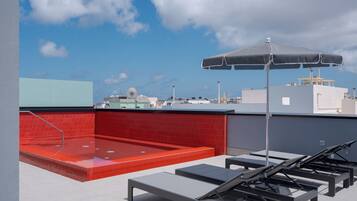 Terraza en la azotea con suelo de tejas rojas, jacuzzi y sillones de descanso bajo un gran paraguas.