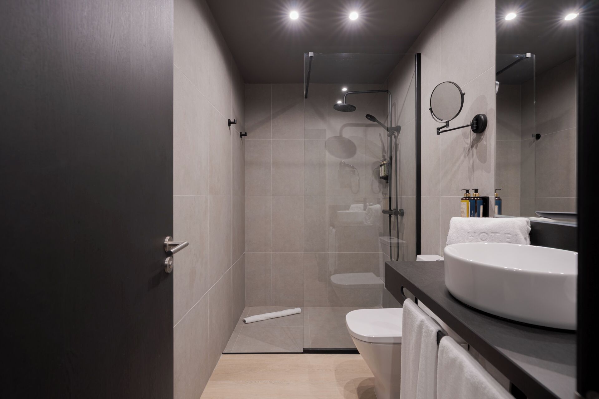 Un baño moderno con ducha envuelta en vidrio, un espejo redondo y un lavamanos blanco con una toalla.