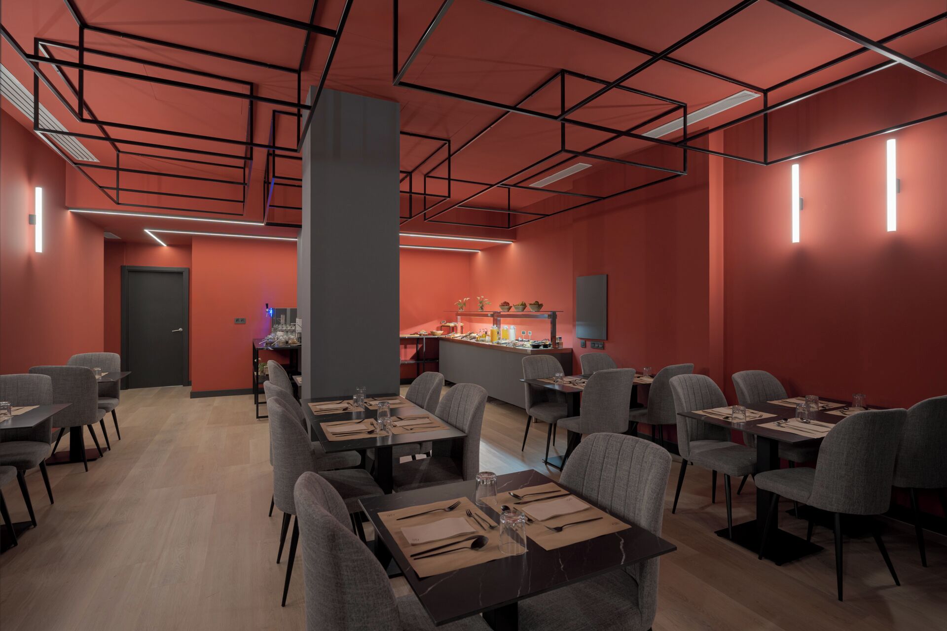A modern dining area with rectangular tables and grey chairs, a central pillar, and a counter with food items.