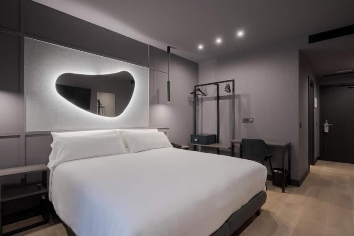 A modern hotel room with a large bed, a desk, a mirror, and a wardrobe.