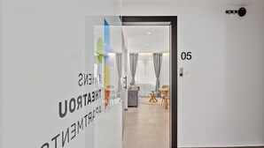 A modern hotel corridor with a glass wall displaying 'THEATER' and 'ENTERTAINMENT' signs, a number '05' on the wall, and a view into a room with a sofa, curtains, and a dining area. - Athens Theatrou Apartments (Athens)