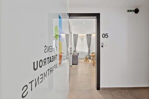 A modern hotel corridor with a glass wall displaying 'THEATER' and 'ENTERTAINMENT' signs, a number '05' on the wall, and a view into a room with a sofa, curtains, and a dining area. - Athens Theatrou Apartments (Athens)
