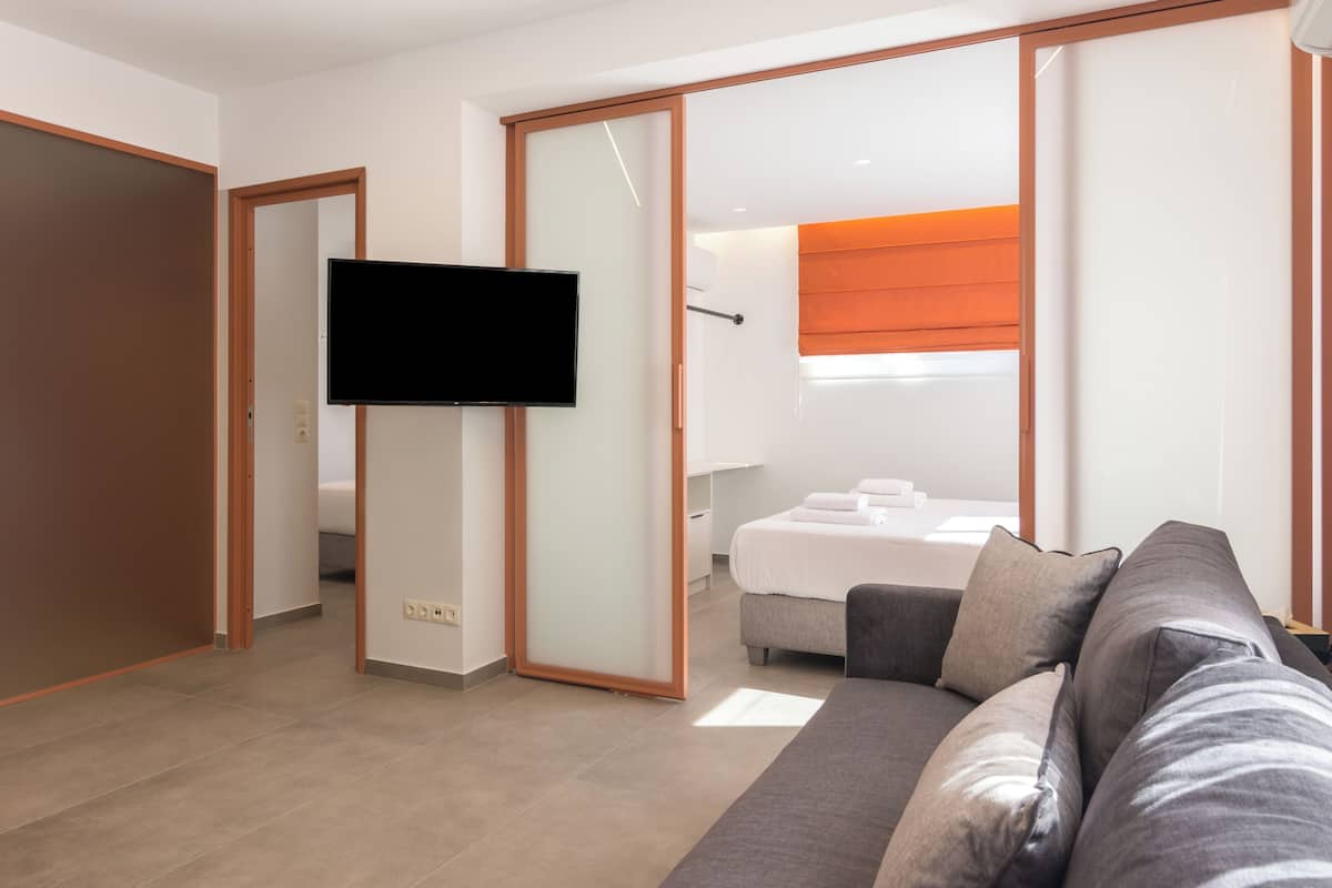 A modern hotel room with a flat-screen TV mounted on the wall, a sofa, a bed with a white headboard, and a sliding glass door leading to a bathroom.