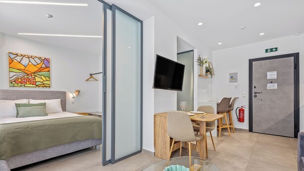 A modern hotel room with a bed, a dining area, a TV, and a door with an emergency exit sign. - Athens Theatrou Apartments (Athens)