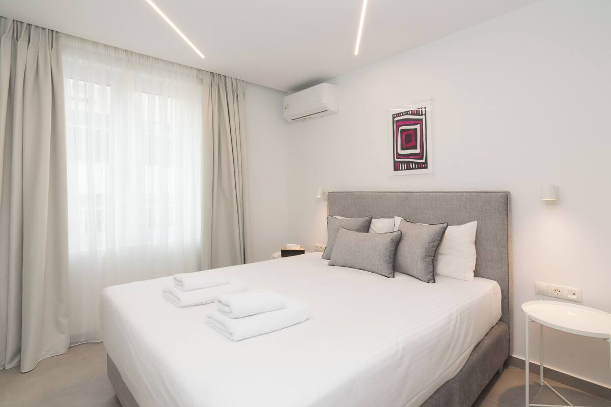 A modern hotel room with a large bed, a bedside table, an air conditioning unit, and a wall-mounted artwork.