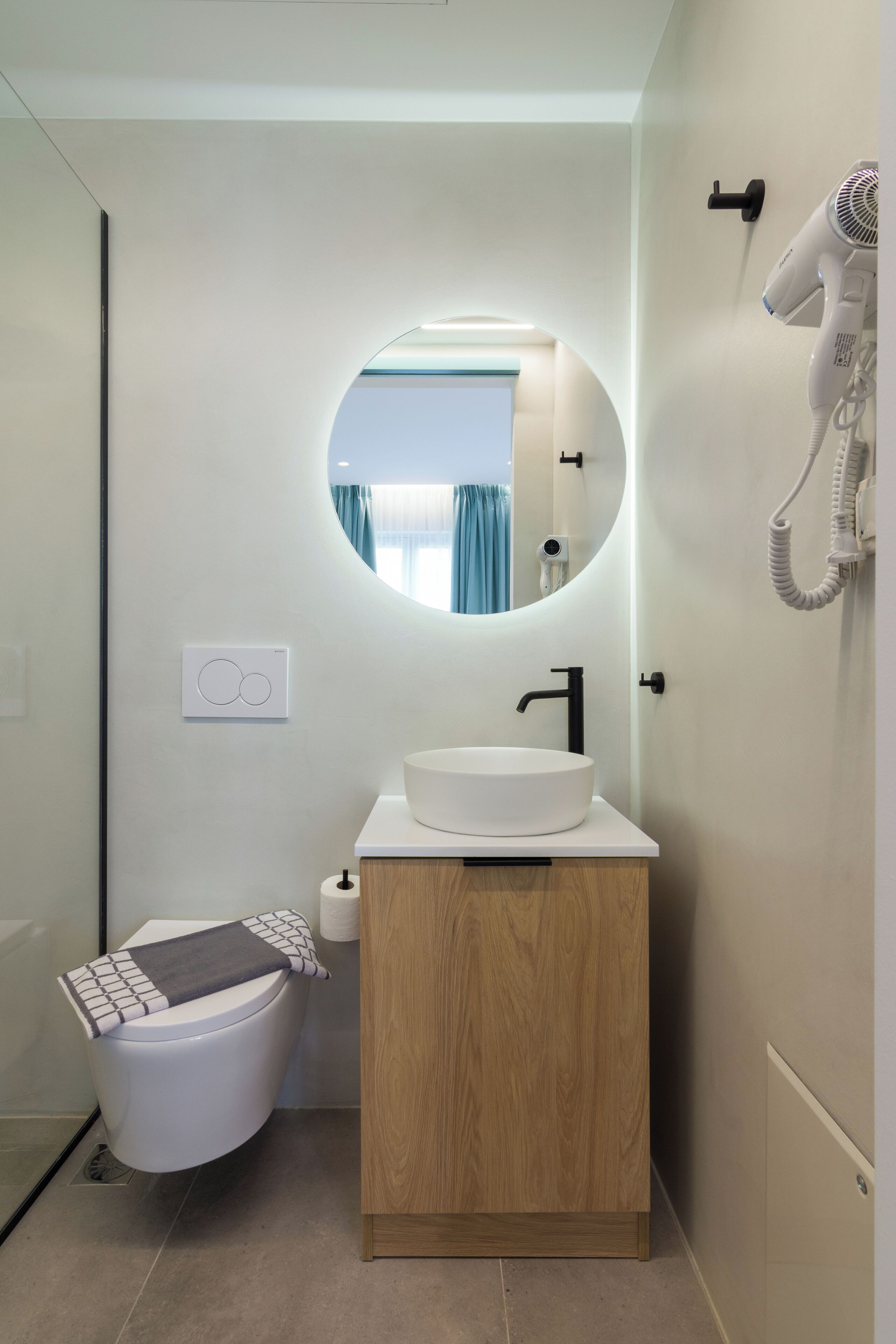 A modern bathroom with a round mirror, a white sink, and a hairdryer mounted on the wall.