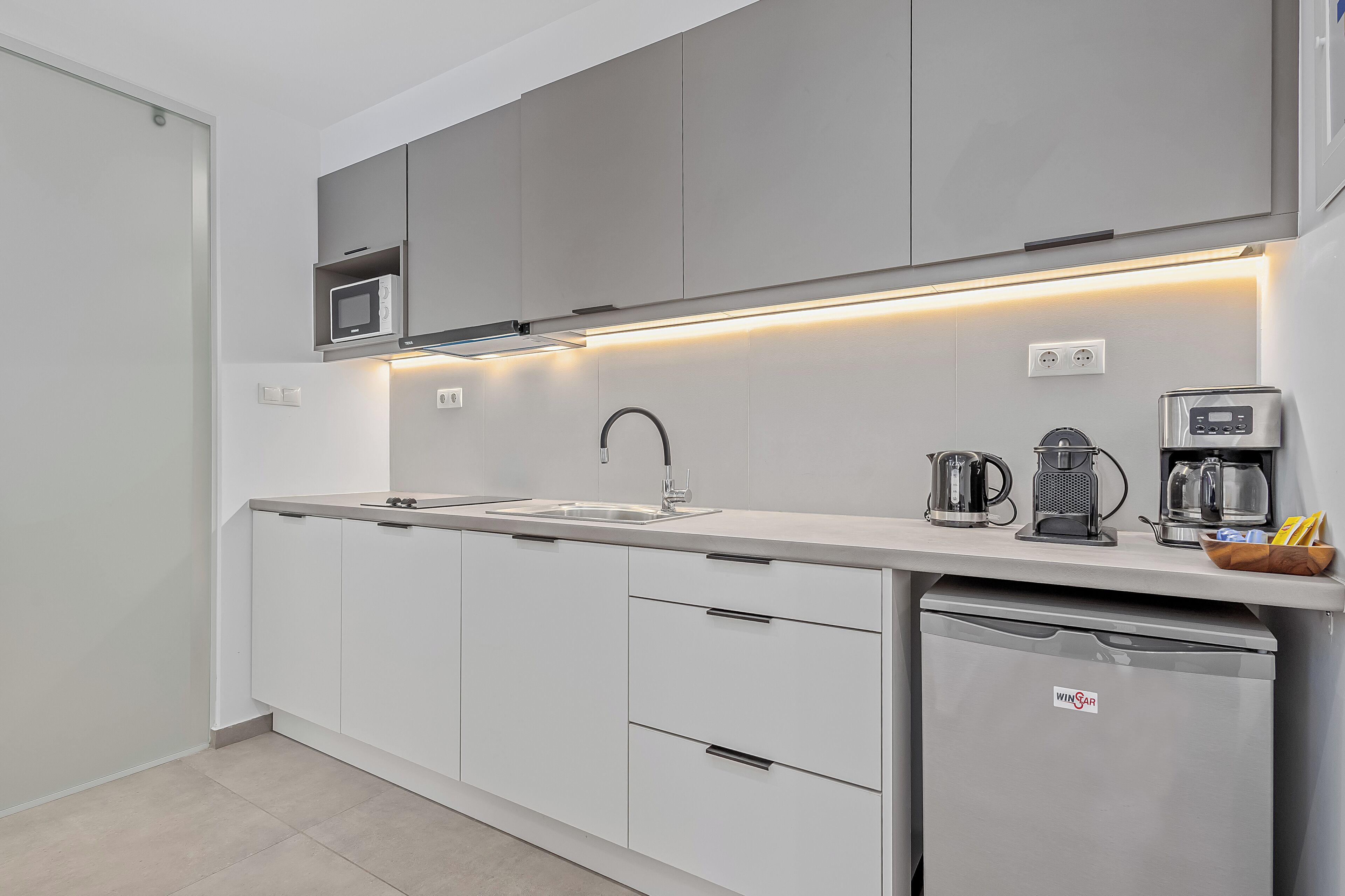 A modern kitchen with white cabinetry, a stainless steel sink, a microwave, a kettle, and a coffee maker.