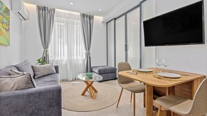 A modern living room with a sofa, a glass-top coffee table, a dining table with chairs, and a flat-screen TV mounted on the wall. - Athens Theatrou Apartments (Athens)