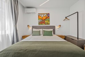 A neatly made bed with a green blanket, a wall-mounted air conditioner, a colorful painting, and a bedside lamp. - Athens Theatrou Apartments (Athens)