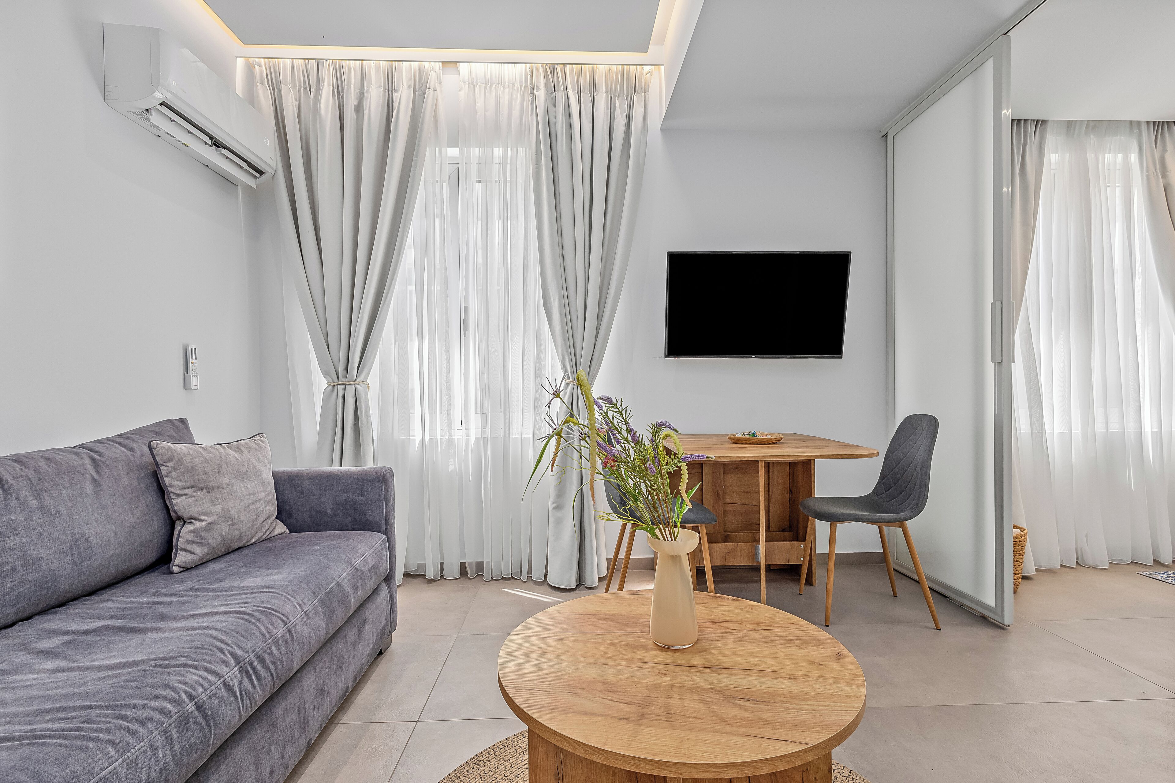 A modern living room with a grey sofa, a wooden coffee table, a dining table with chairs, and a flat-screen TV mounted on the wall.