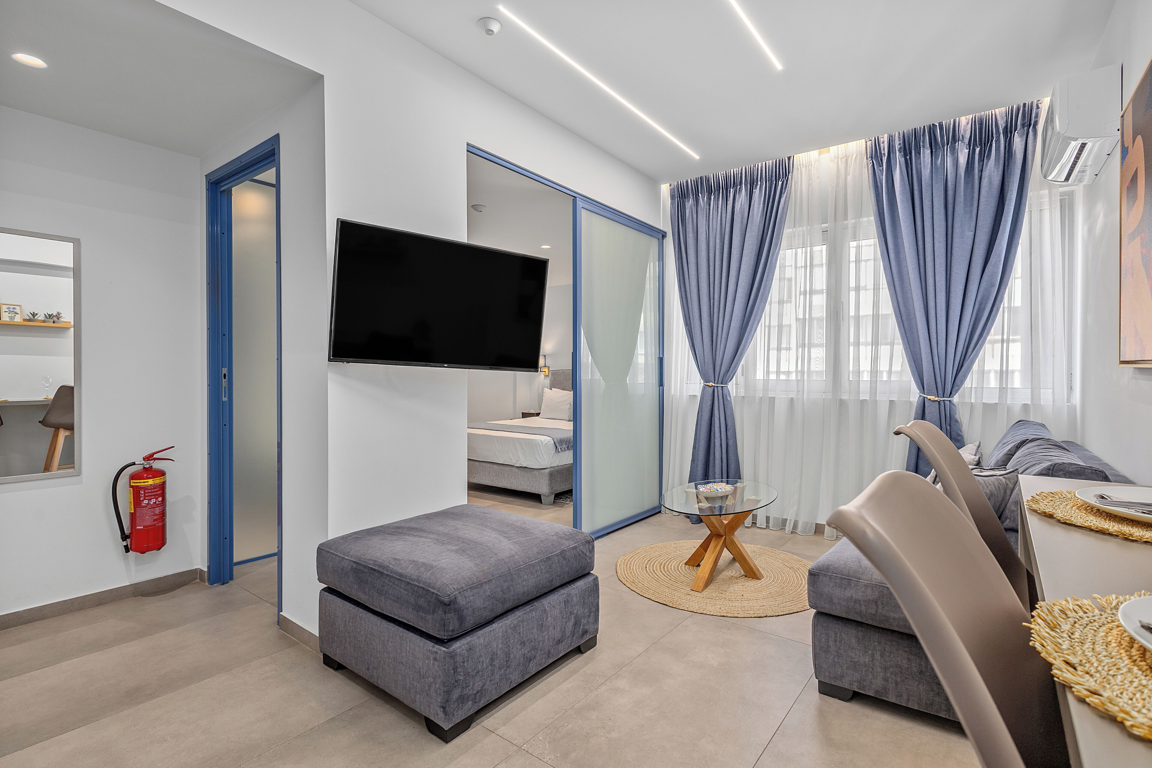 A modern living room with a flat-screen TV, grey sofas, a glass coffee table, and a fire extinguisher on the wall.