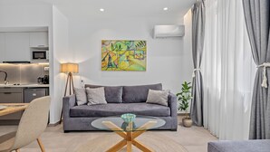 A modern living room with a grey sofa, a glass coffee table, a dining area with a wooden table and chairs, and a kitchen in the background. - Athens Theatrou Apartments (Athens)