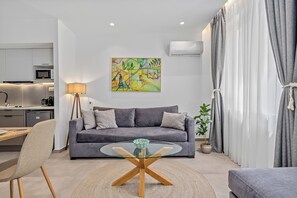 A modern living room with a grey sofa, a glass coffee table, a dining area with a wooden table and chairs, and a kitchen in the background.