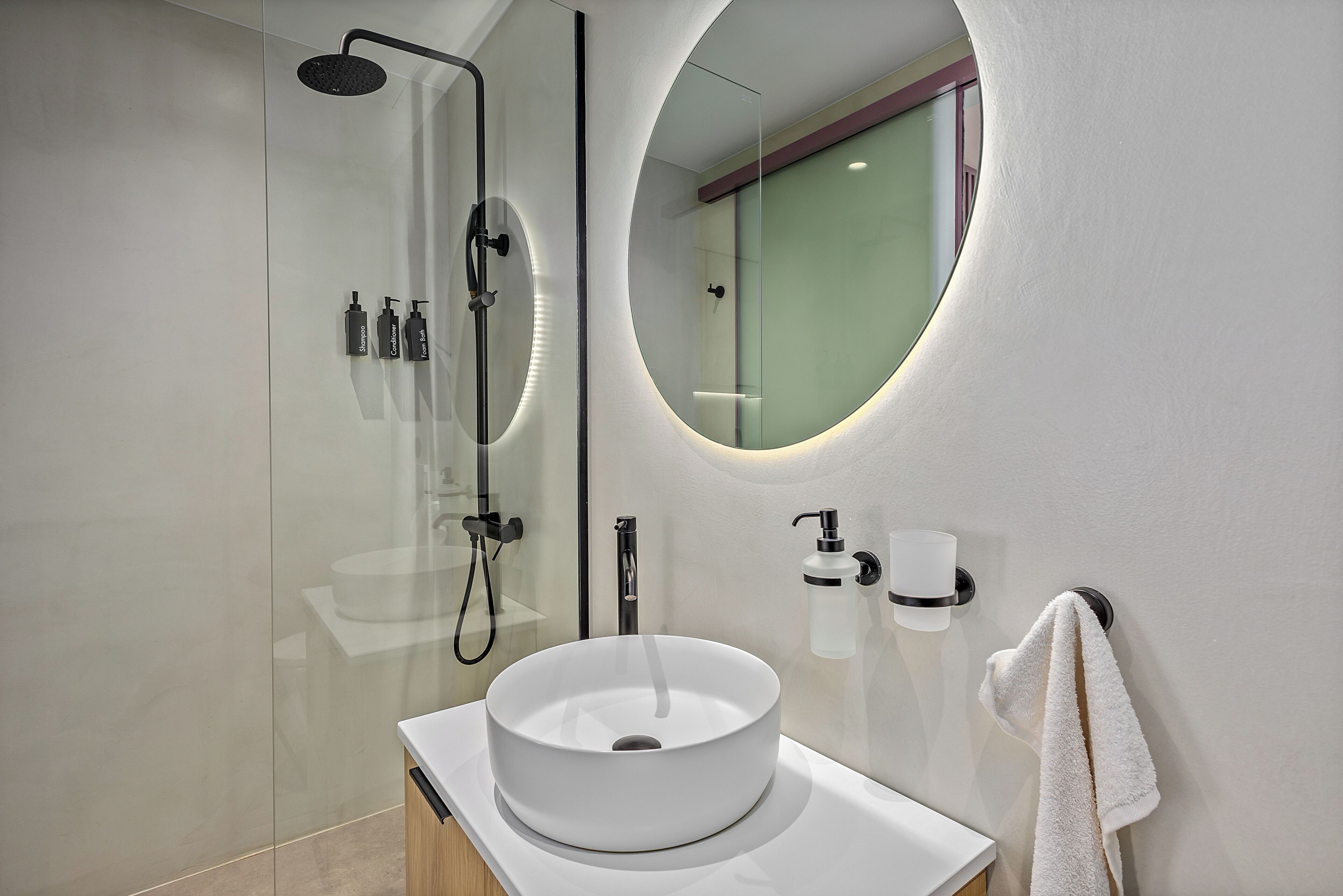 A modern bathroom with a glass-enclosed shower, a round mirror, a white sink, and a towel hanging on the wall.