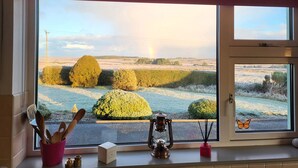 A kitchen with a large window overlooking a rural landscape, a clock on the wall, and kitchen utensils on the windowsill.