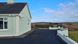 A house with a paved driveway leading to a gate, surrounded by a grassy area and a view of distant fields.