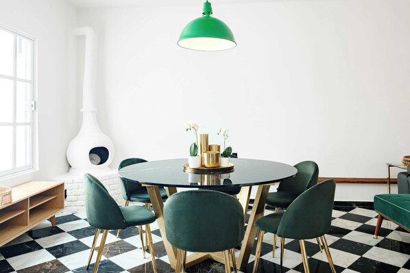 A dining area with a round table, green chairs, a green pendant light, and a white wall with a window.